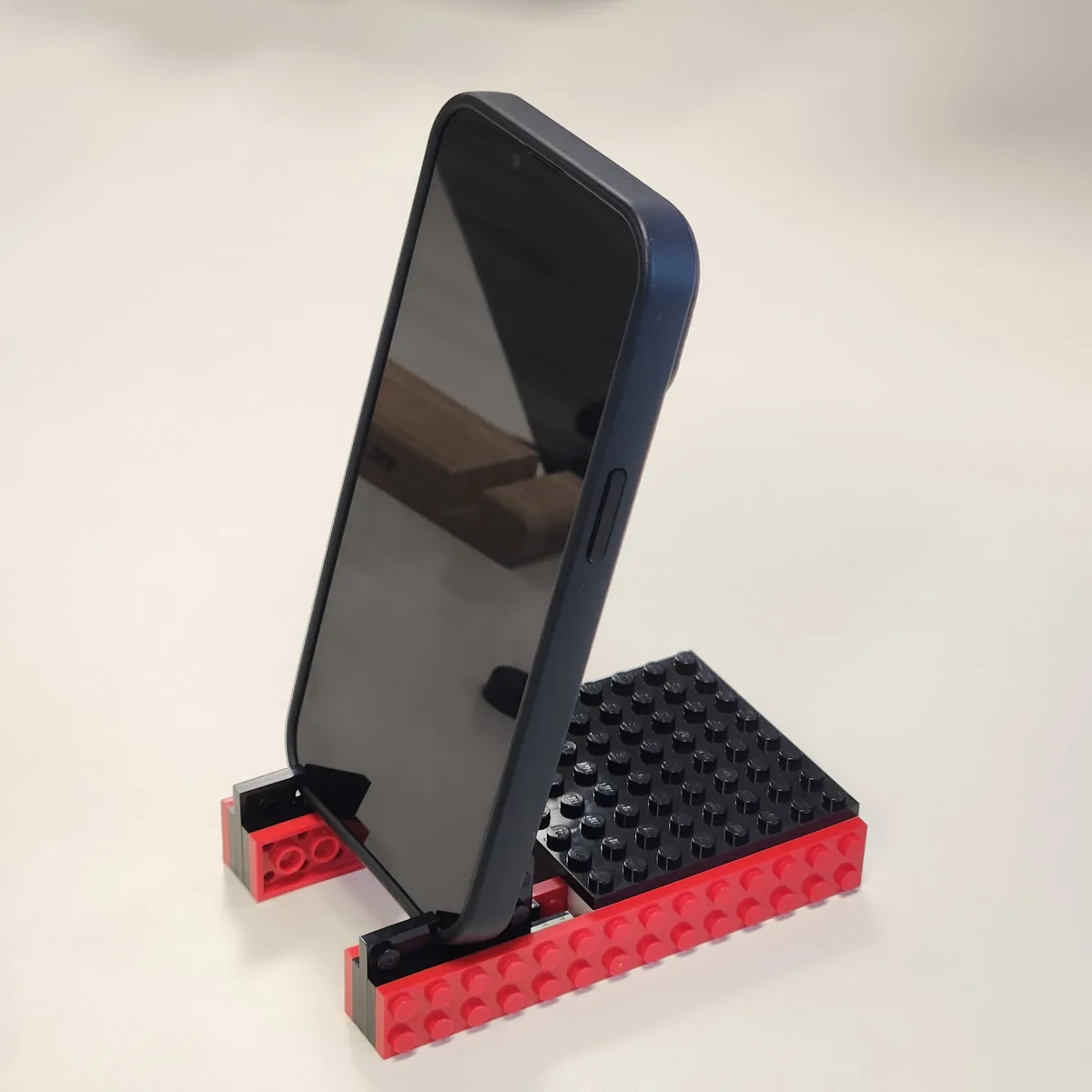 A smartphone with a dark blue case is propped upright on a makeshift stand constructed from red and black Lego bricks. The…
