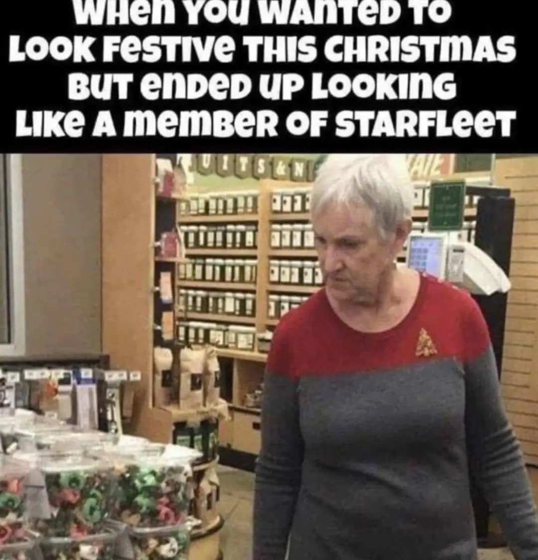 An older woman with short white hair stands in a store aisle, wearing a red-and-grey sweater with a Starfleet insignia pin…