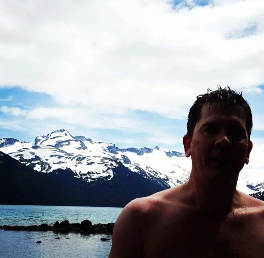 A shirtless man with wet hair stands in water, smiling at the camera, with a backdrop of snow-capped mountains and a partly…
