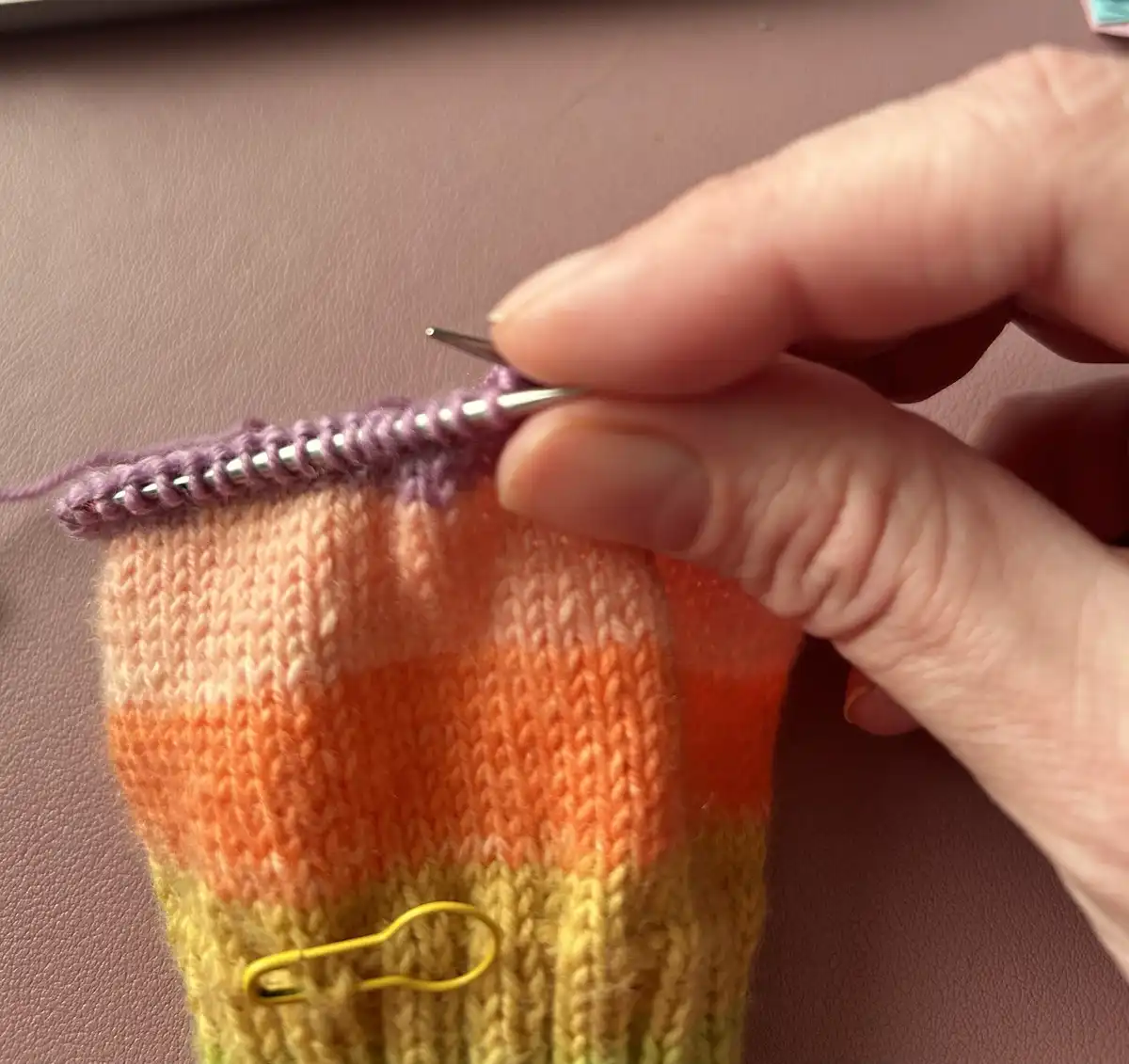 A close-up photo shows a hand holding knitting needles while working on a colorful, striped sock in progress. The sock…