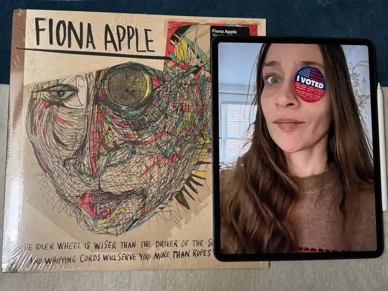A person with an 'I VOTED' sticker over one eye is shown on a tablet screen, placed next to a physical copy of Fiona Apple's…