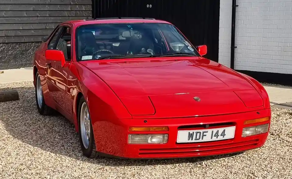 A glossy red Porsche 944 sports car is parked on gravel in front of a black and white brick wall. The car's license plate…