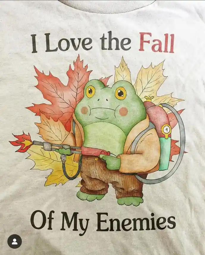 A t-shirt graphic shows a cartoon green frog wearing a brown jacket and pants, holding a flamethrower with a flame, and…