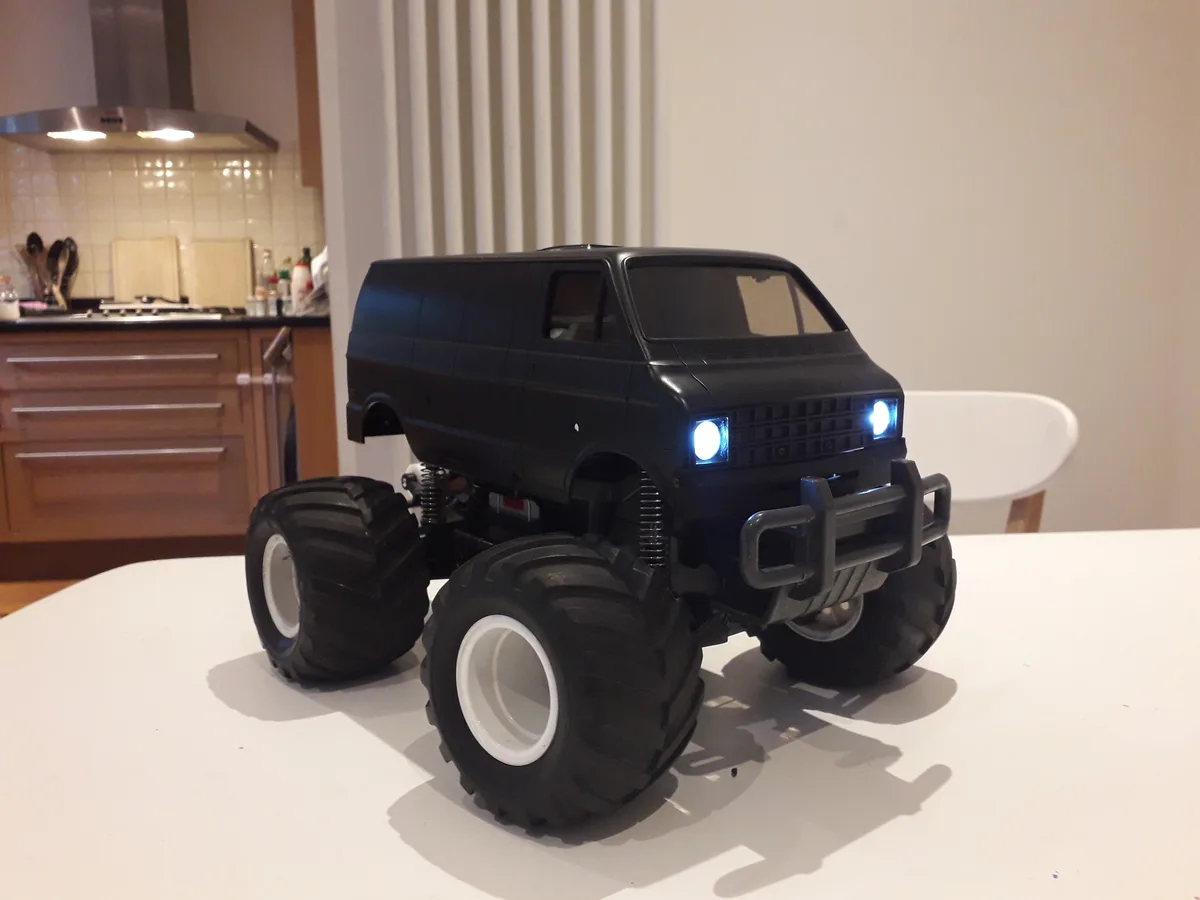 A black toy monster truck van with oversized tires and glowing headlights sits on a white kitchen table, with a kitchen…