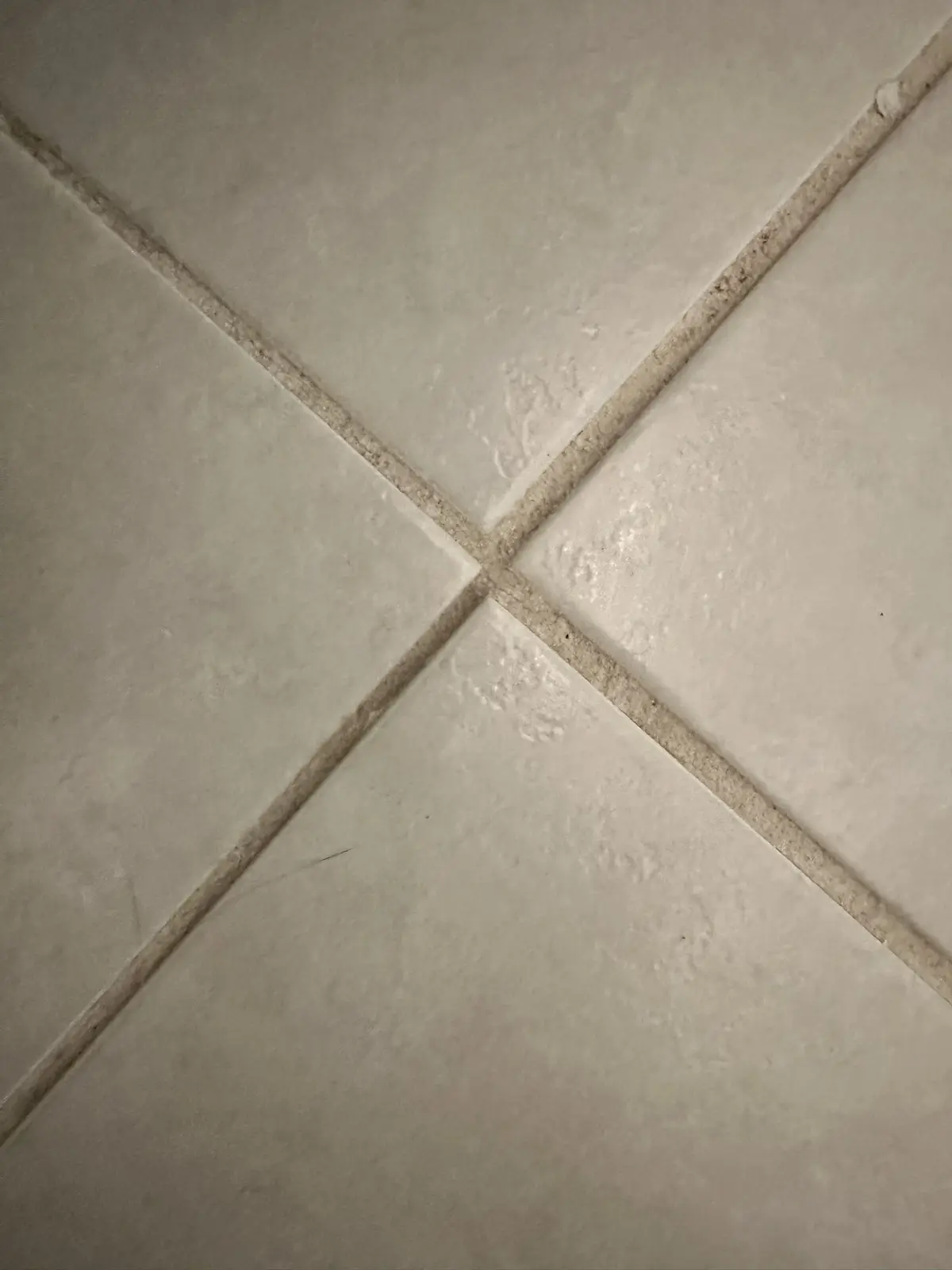 A close-up photo of four light-colored square tiles arranged in a diamond pattern, with dirty, discolored grout lines…