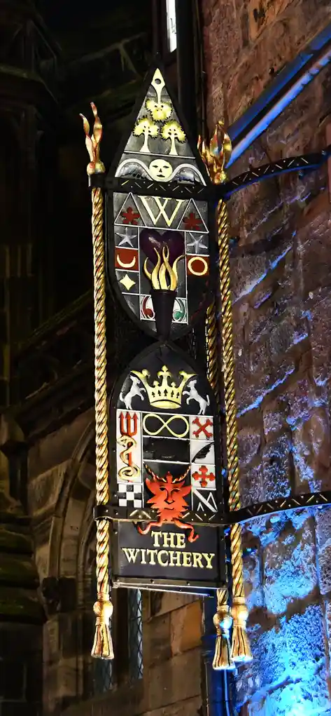 A vertically oriented, ornate, black metal sign with gold and colored embellishments hangs against a stone wall. The sign…
