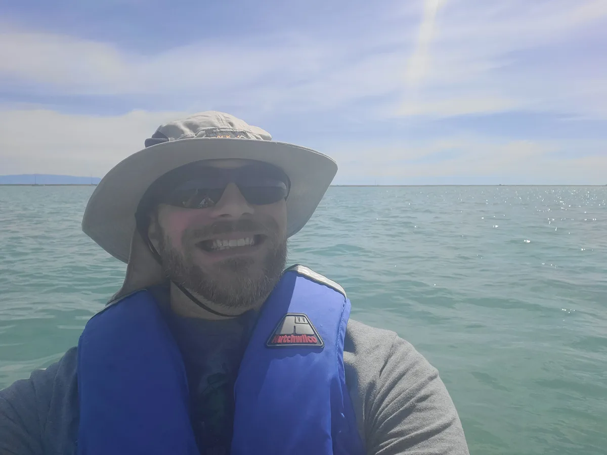 A man with a beard, wearing a wide-brimmed hat, sunglasses, and a blue life vest, smiles broadly while taking a selfie on a…