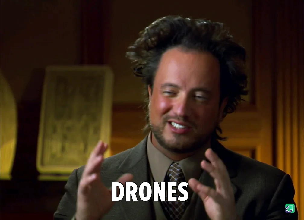 A man with curly dark hair and a beard, wearing a suit and tie, smiles while gesturing with both hands. The word 'DRONES' is…