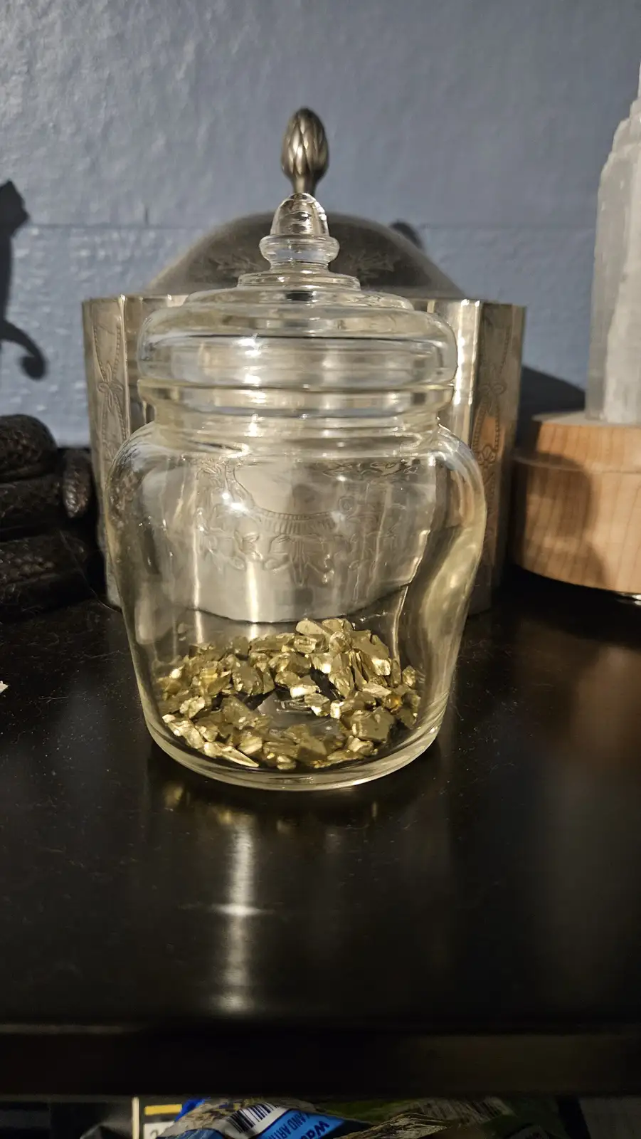 A clear glass jar with a silver lid sits on a dark surface, containing only a small amount of shiny, irregular gold-colored…
