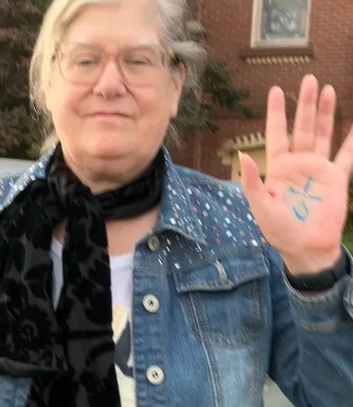 an older woman with glasses and a denim jacket holds up her left hand, which has a blue 'no' symbol drawn on the palm, while…