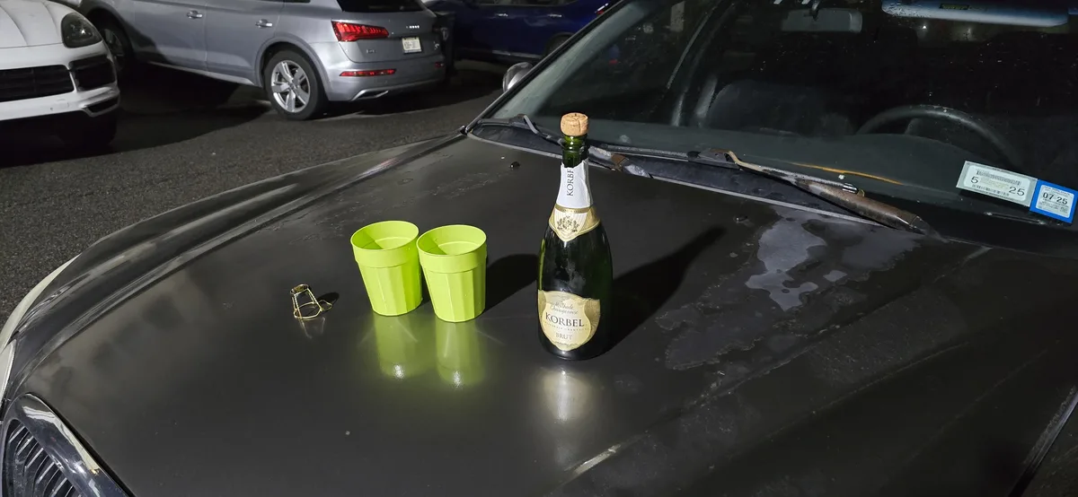 A bottle of Korbel Brut champagne and two bright green plastic cups sit on the hood of a black car in a parking lot at…