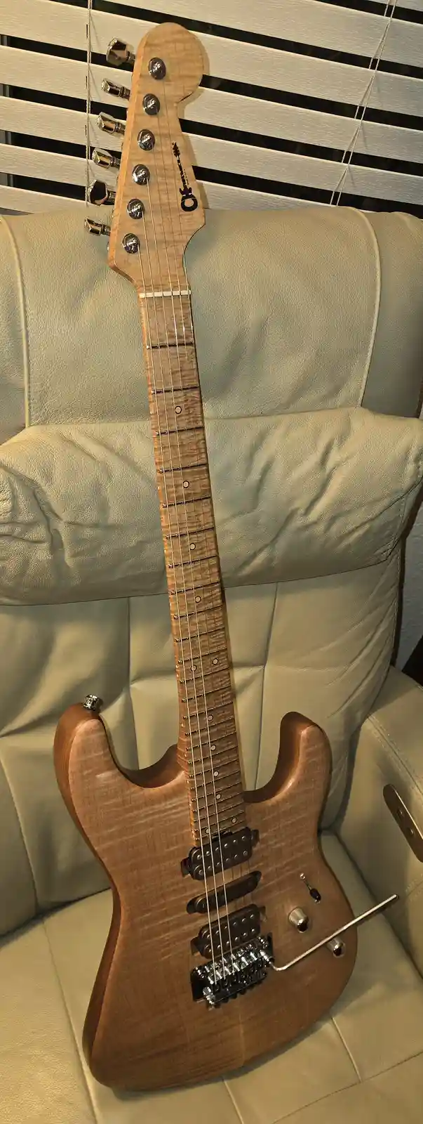 A light-colored electric guitar with a visible wood grain finish is propped upright against the back of a beige leather…