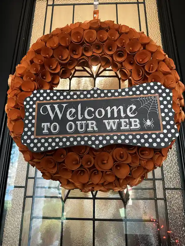 An orange Halloween wreath made of rolled paper hangs on a glass door with a black frame. A black sign with white polka dots…