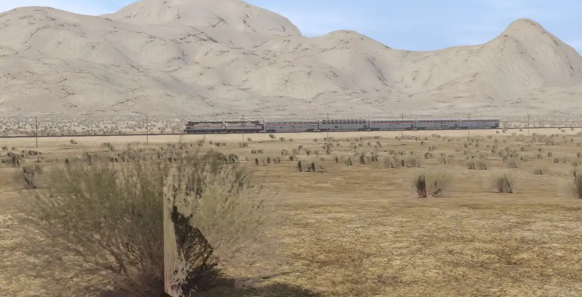 A long silver passenger train travels across a barren desert landscape with sandy hills in the background. Sparse shrubs dot…