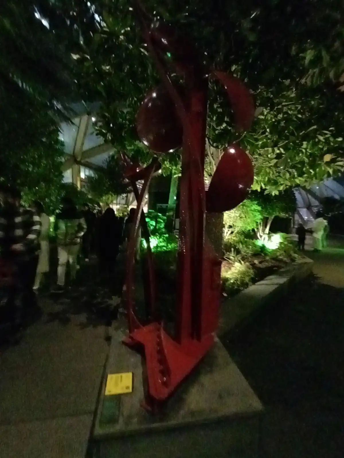 A nighttime photo of a red, abstract metal sculpture resembling a stylized tree with rounded, bulbous shapes instead of…