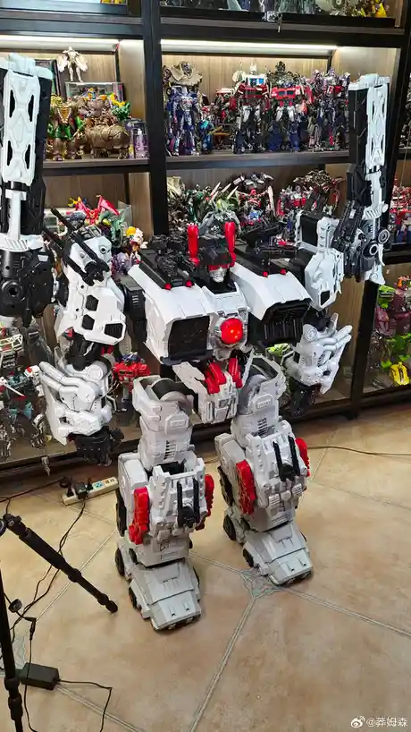 A large, white and red robot toy stands in a room with shelves filled with other Transformers action figures. The robot has…