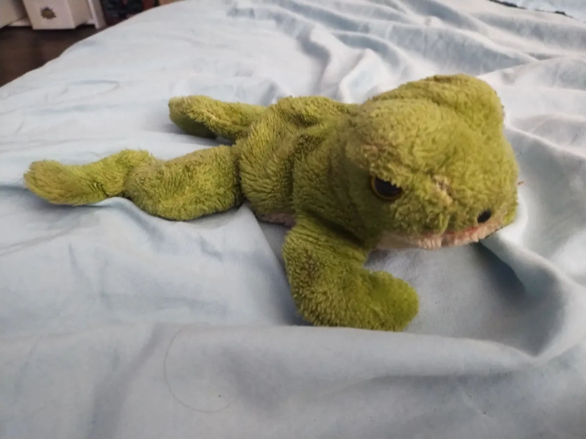 A worn, green, fuzzy stuffed animal resembling a lizard lies on a rumpled light blue bedsheet. The toy has one visible dark…