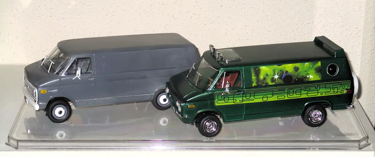 Two model vans sit side-by-side on a clear display stand: a plain gray van and a green van with a space-themed decal showing…