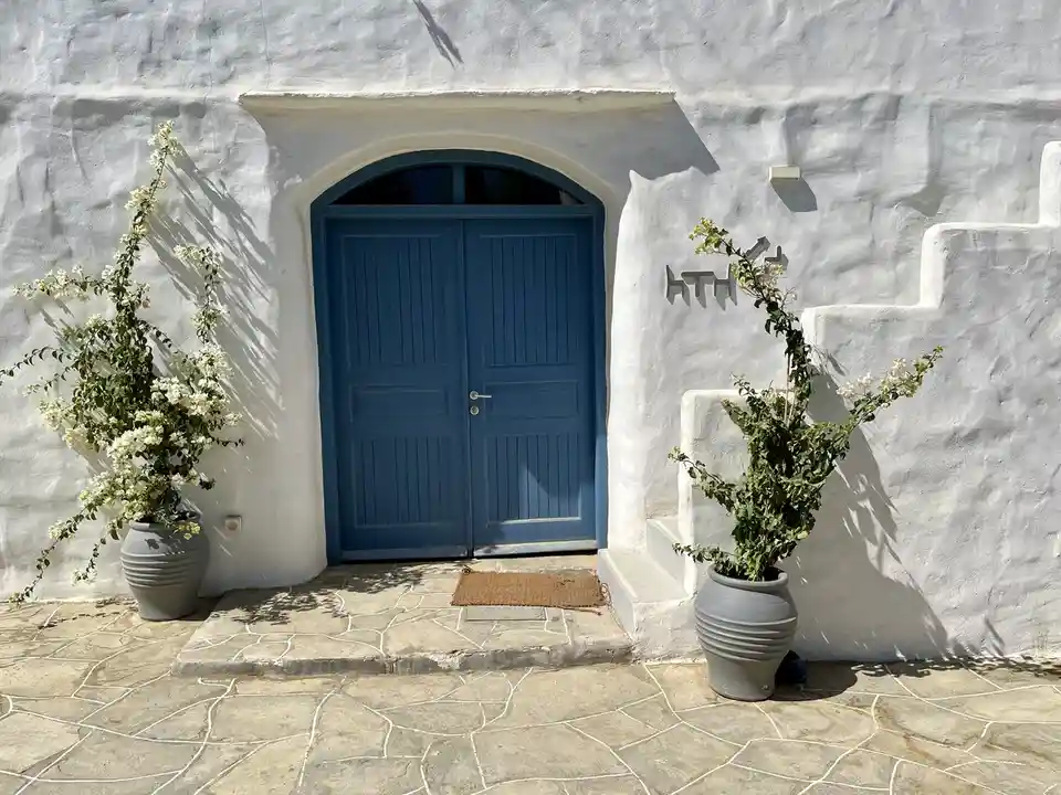 A picturesque white-washed building with a blue arched door, flanked by two large grey pots with blooming white flowers. The…