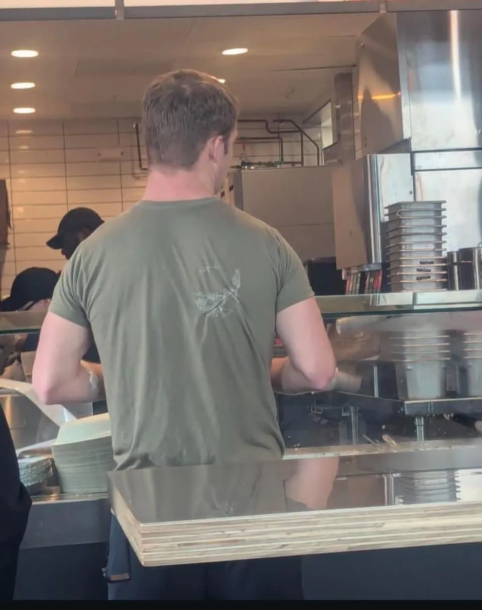A man in a green t-shirt is seen from behind, working behind a glass counter in a coffee shop. His shirt has a faded…