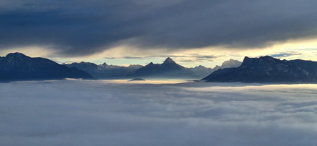 A panoramic view of majestic, snow-capped mountains rising above a dense, sea-like layer of clouds, with a dramatic sky…