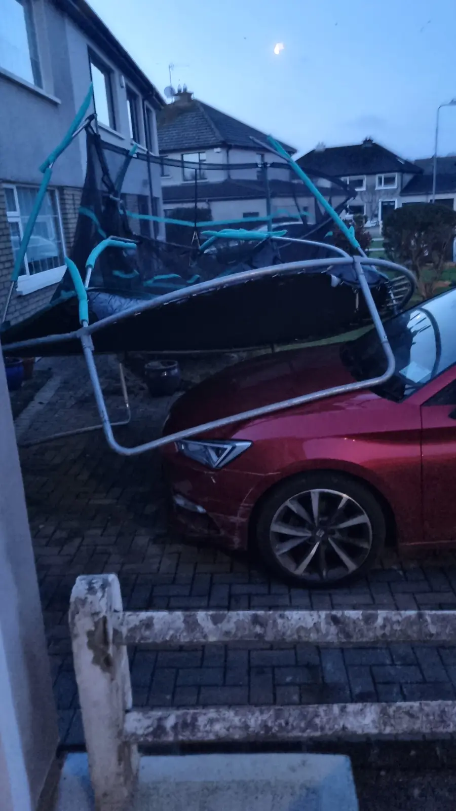 A red car is parked on a paved driveway with a trampoline collapsed and draped over its roof and hood, its metal frame and…