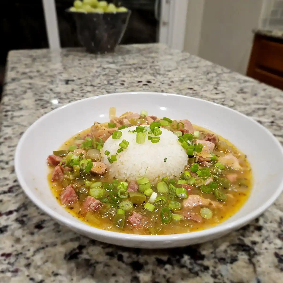 A white bowl of jambalaya with a mound of white rice in the center, topped with chopped green onions, sits on a speckled…