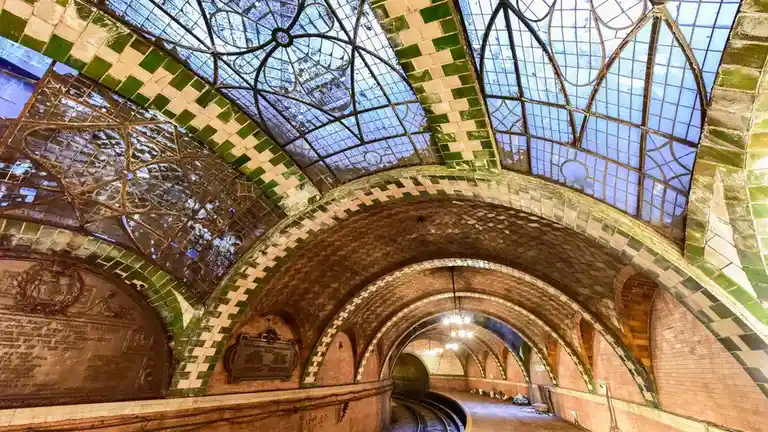 a photograph of an ornate, arched subway station with a glass and metal ceiling, green and white tiled arches, and brick…