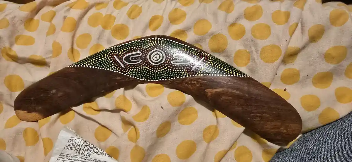 A wooden boomerang with Aboriginal dot art style painting lies on a yellow polka-dot fabric. The word 'cost' is written in…