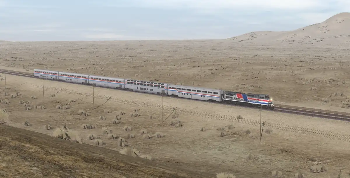 A long Amtrak passenger train travels through a vast, arid desert landscape with sparse vegetation and rolling hills under a…
