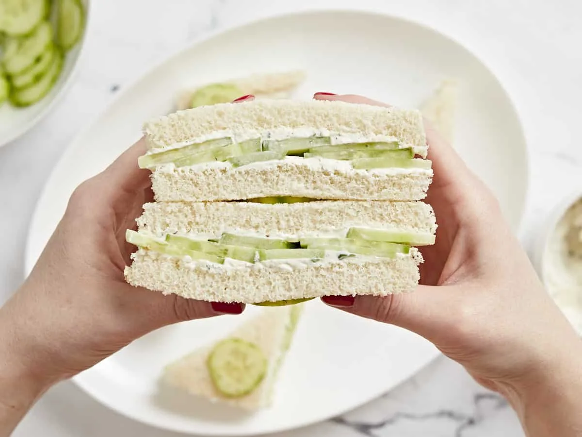 Two hands hold two cucumber sandwiches stacked vertically, revealing layers of cucumber slices and creamy spread between…