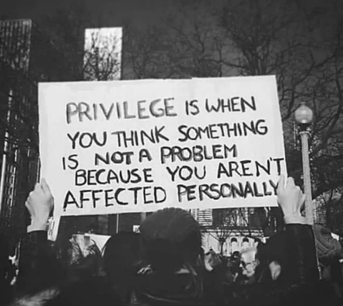 A black-and-white photo shows a person from behind holding a handmade protest sign at night. The sign has handwritten text…
