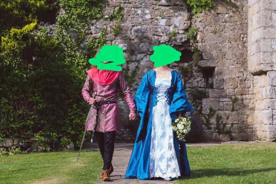 Two people dressed in medieval-style wedding attire walk hand-in-hand along a stone path in front of a stone wall with ivy…