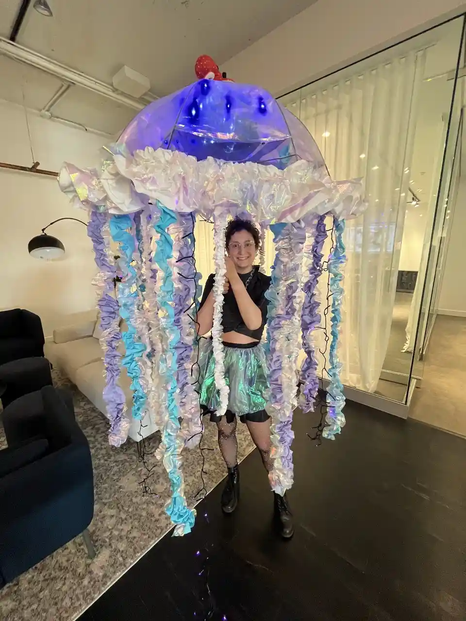 A person stands inside a large, elaborate jellyfish costume made of iridescent fabric in shades of blue, purple, and white…
