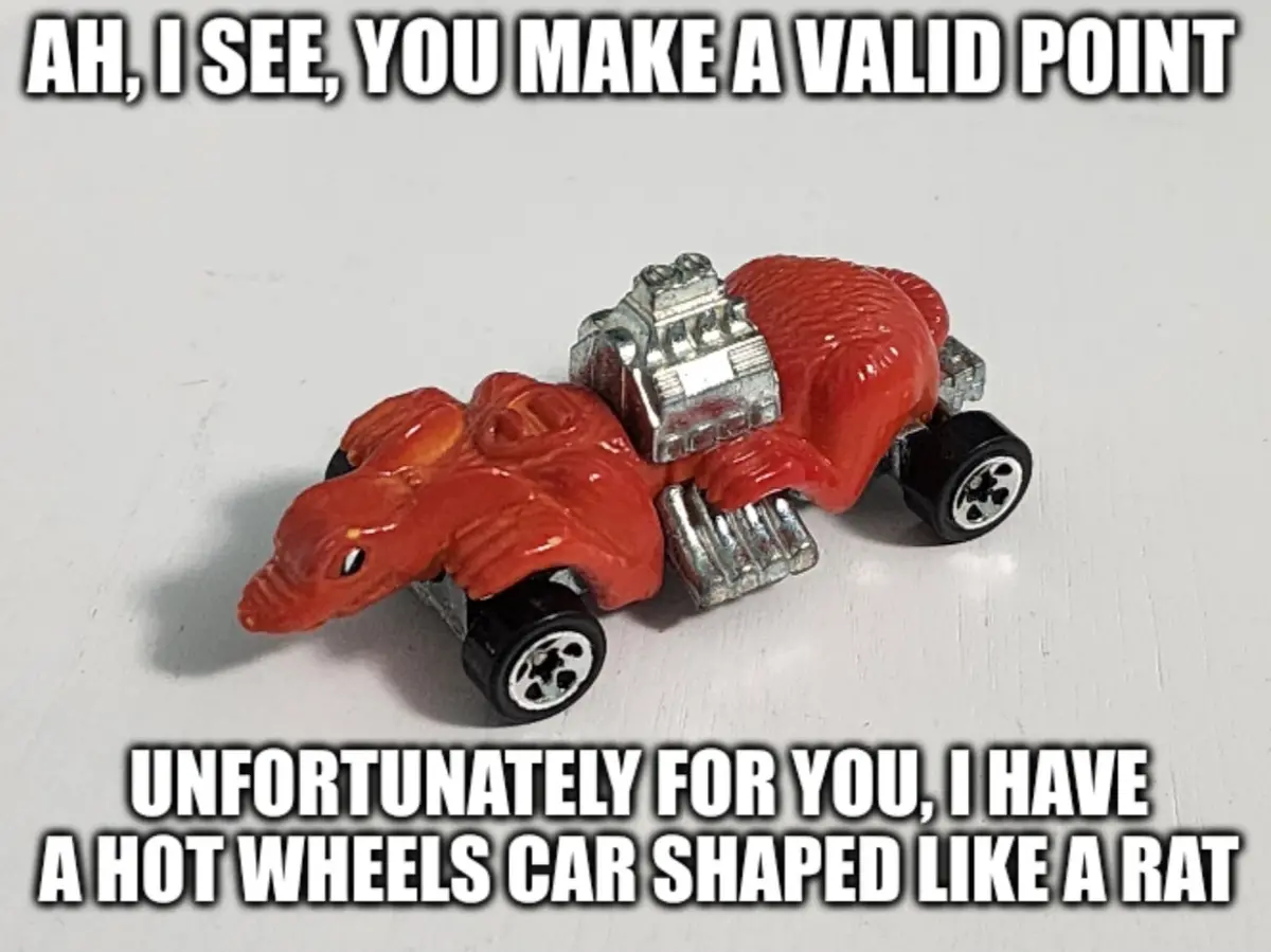 A red Hot Wheels toy car shaped like a rat with a silver engine and black wheels sits on a white surface. The top text reads…