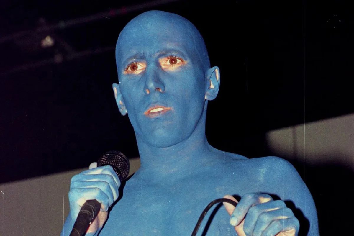 A man with his entire face and upper body painted blue holds a microphone, looking directly at the camera with a slightly…