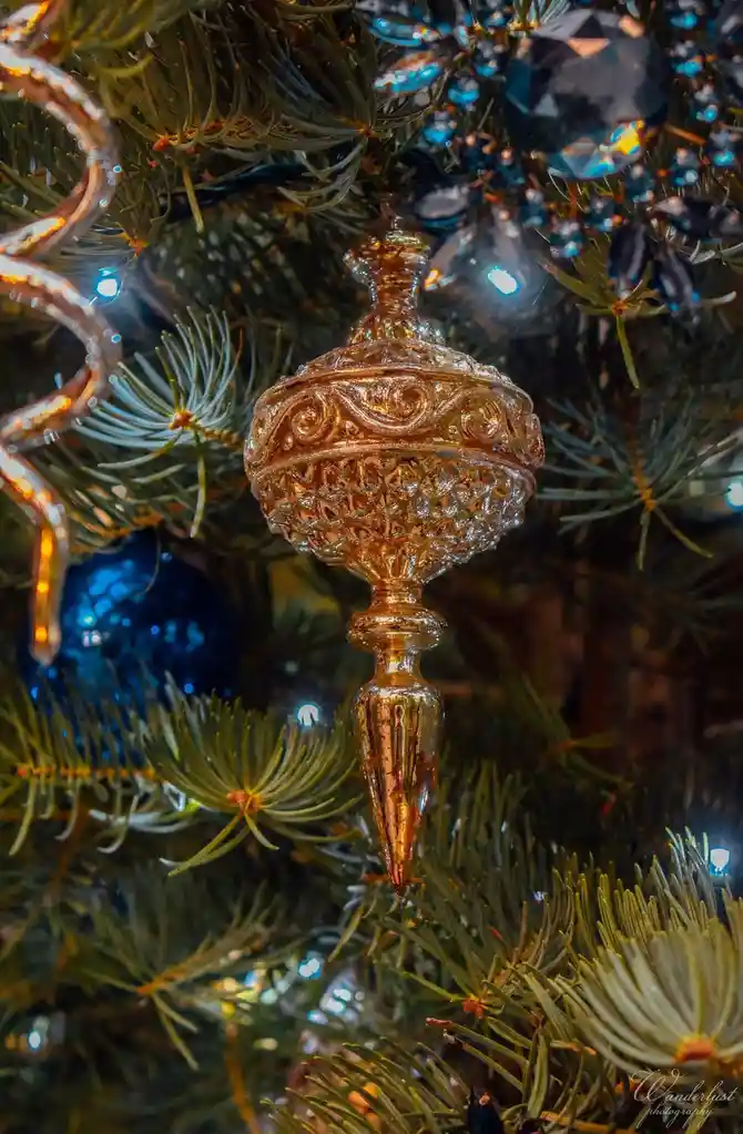 A close-up photo of an ornate, gold-colored glass Christmas ornament hanging on a pine tree, surrounded by other ornaments…