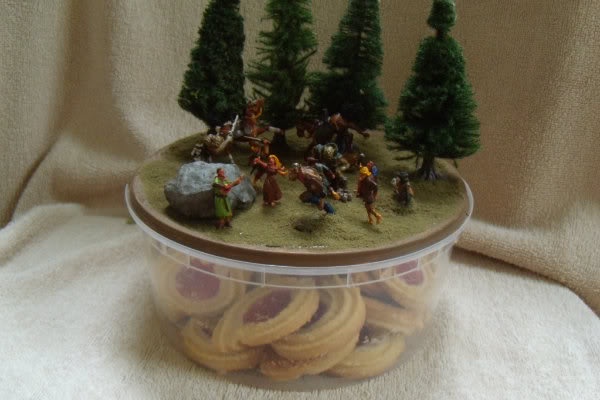 A clear plastic container filled with round, jam-filled cookies has a miniature diorama on its lid, depicting a battle scene…