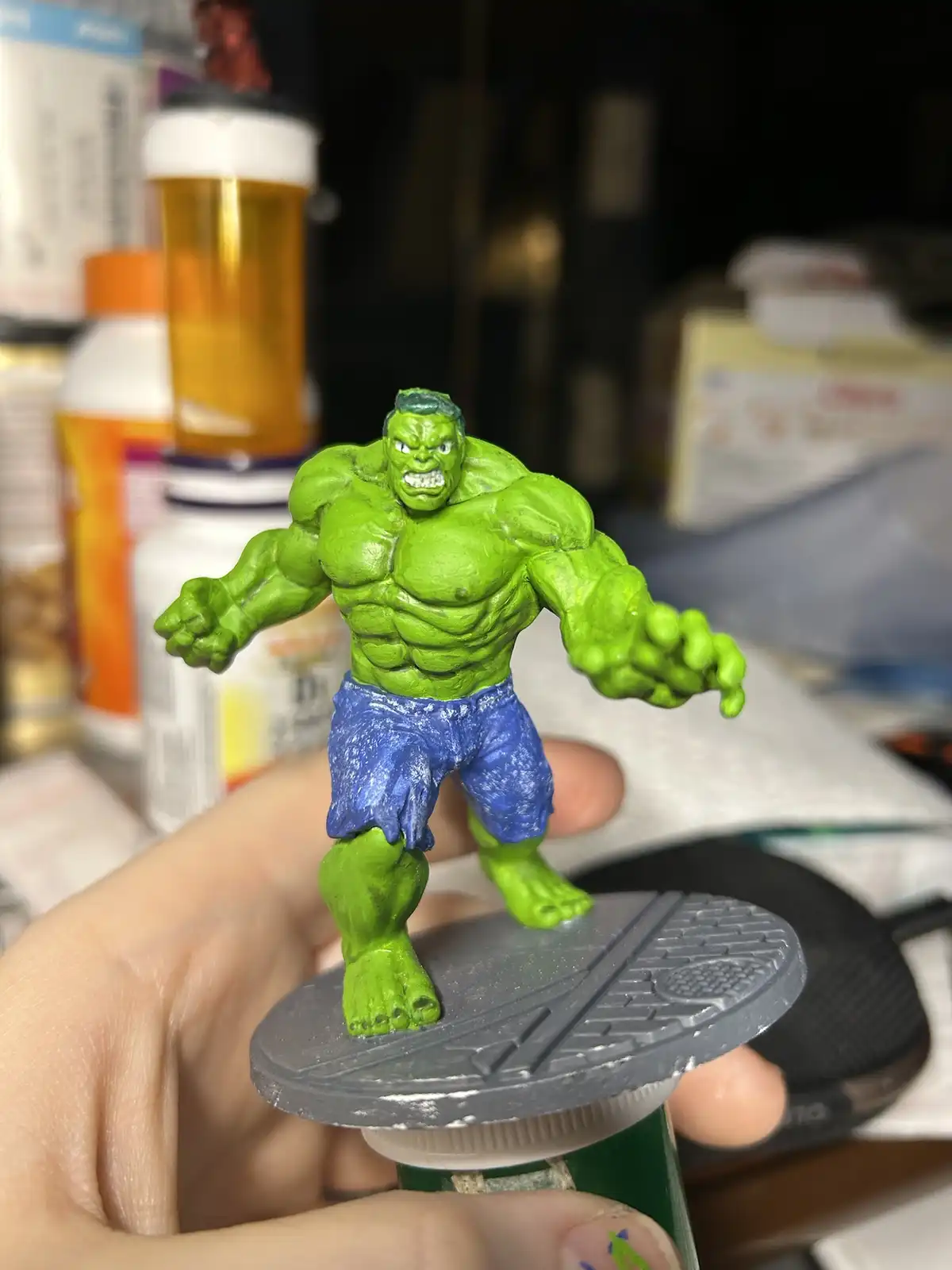 A hand holds a green, muscular Hulk action figure wearing blue shorts, standing on a gray base. The background is cluttered…