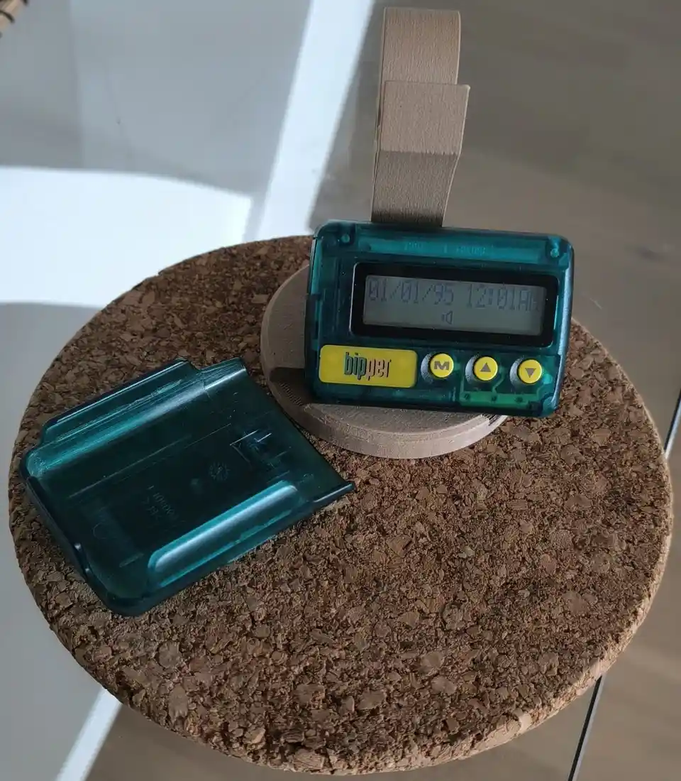 A teal-colored, retro 'bipper' pager with a digital display showing '01/01/95 12:01AM' sits on a cork coaster, next to its…