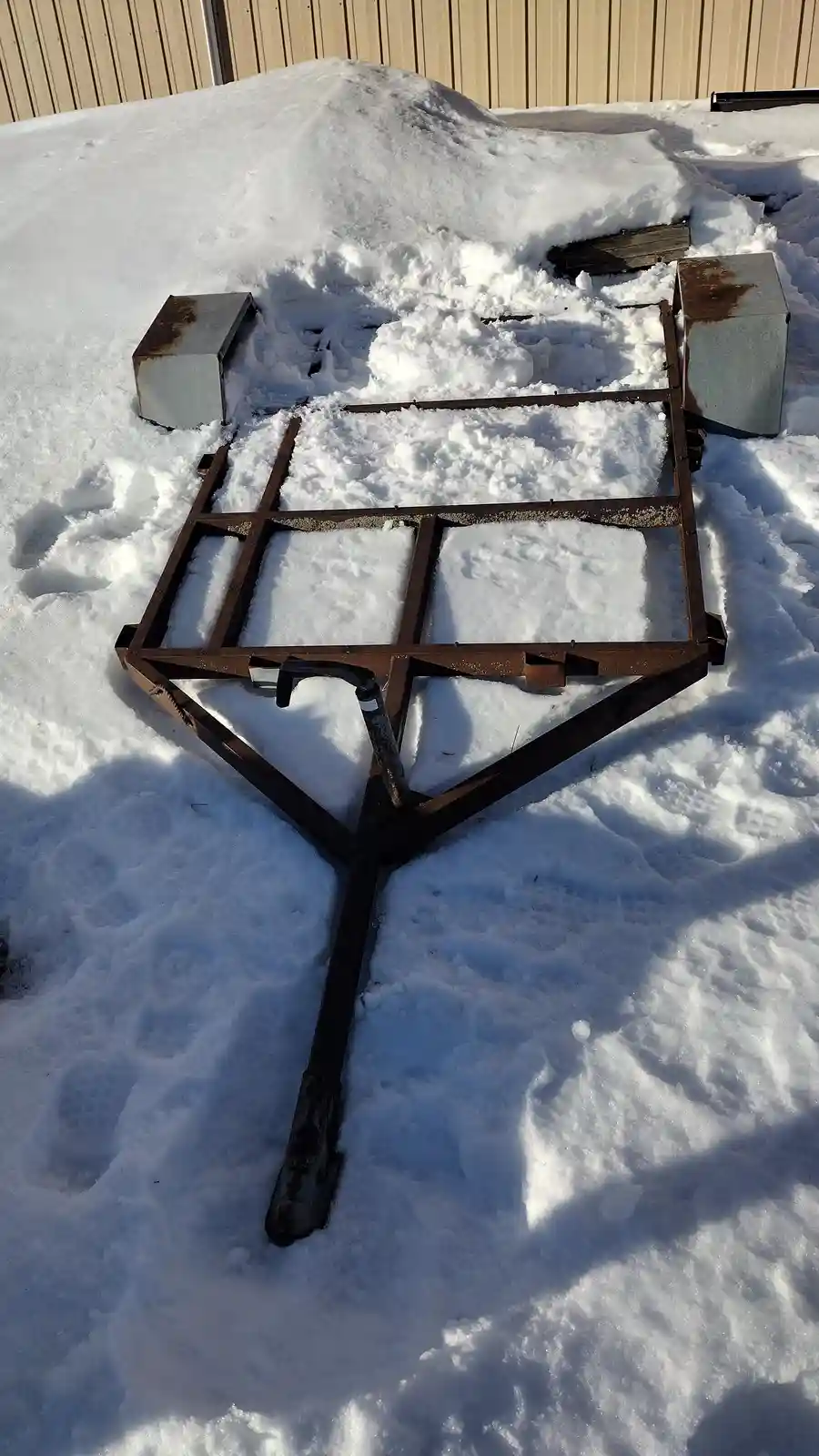 A rusty metal frame, resembling a snow blower or trailer hitch, is partially buried in deep snow outdoors. Two metal boxes…