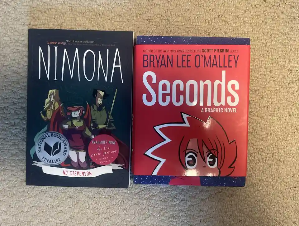 Two graphic novels, 'Nimona' by ND Stevenson and 'Seconds' by Bryan Lee O'Malley, are placed side-by-side on a beige carpet…