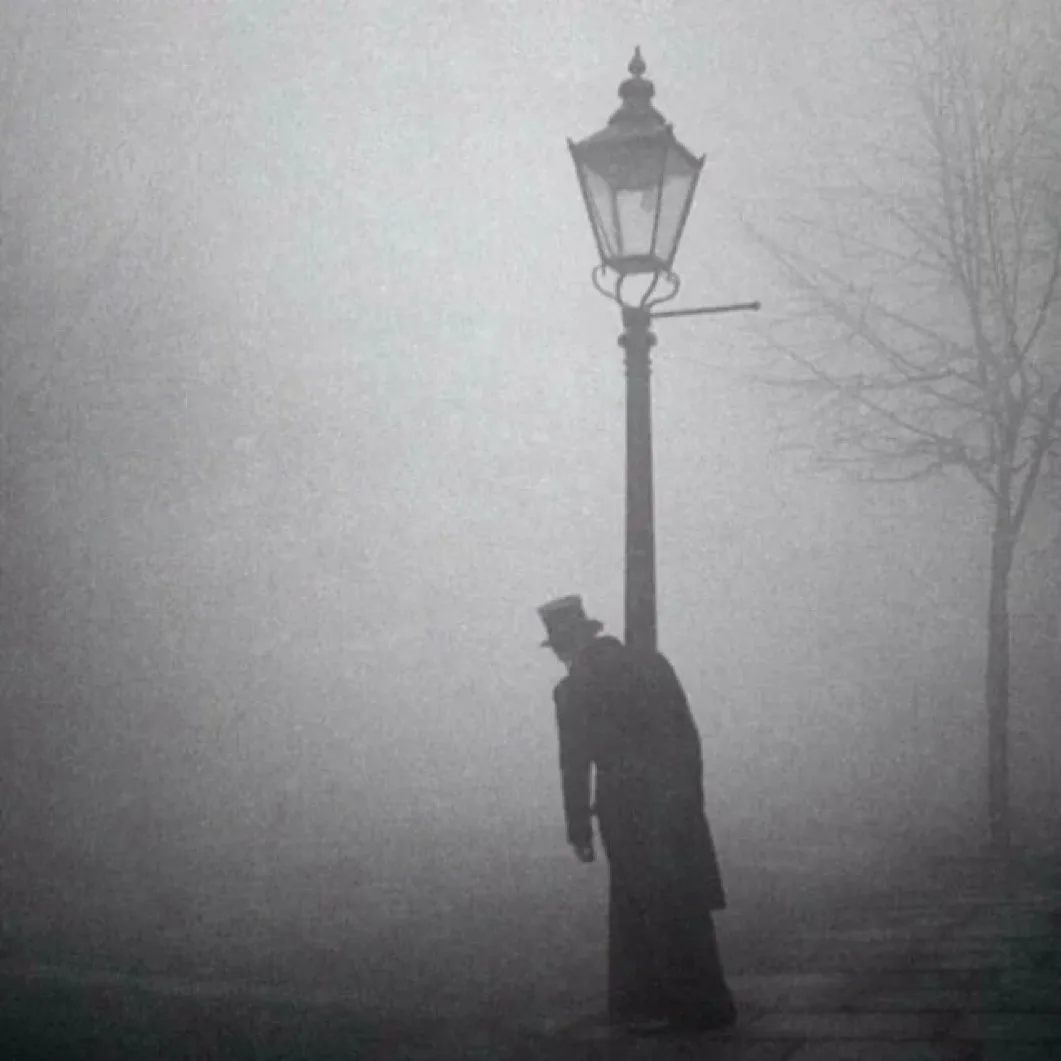 A grayscale, foggy image shows a silhouetted person in a top hat and long coat leaning against a vintage streetlamp. Bare…