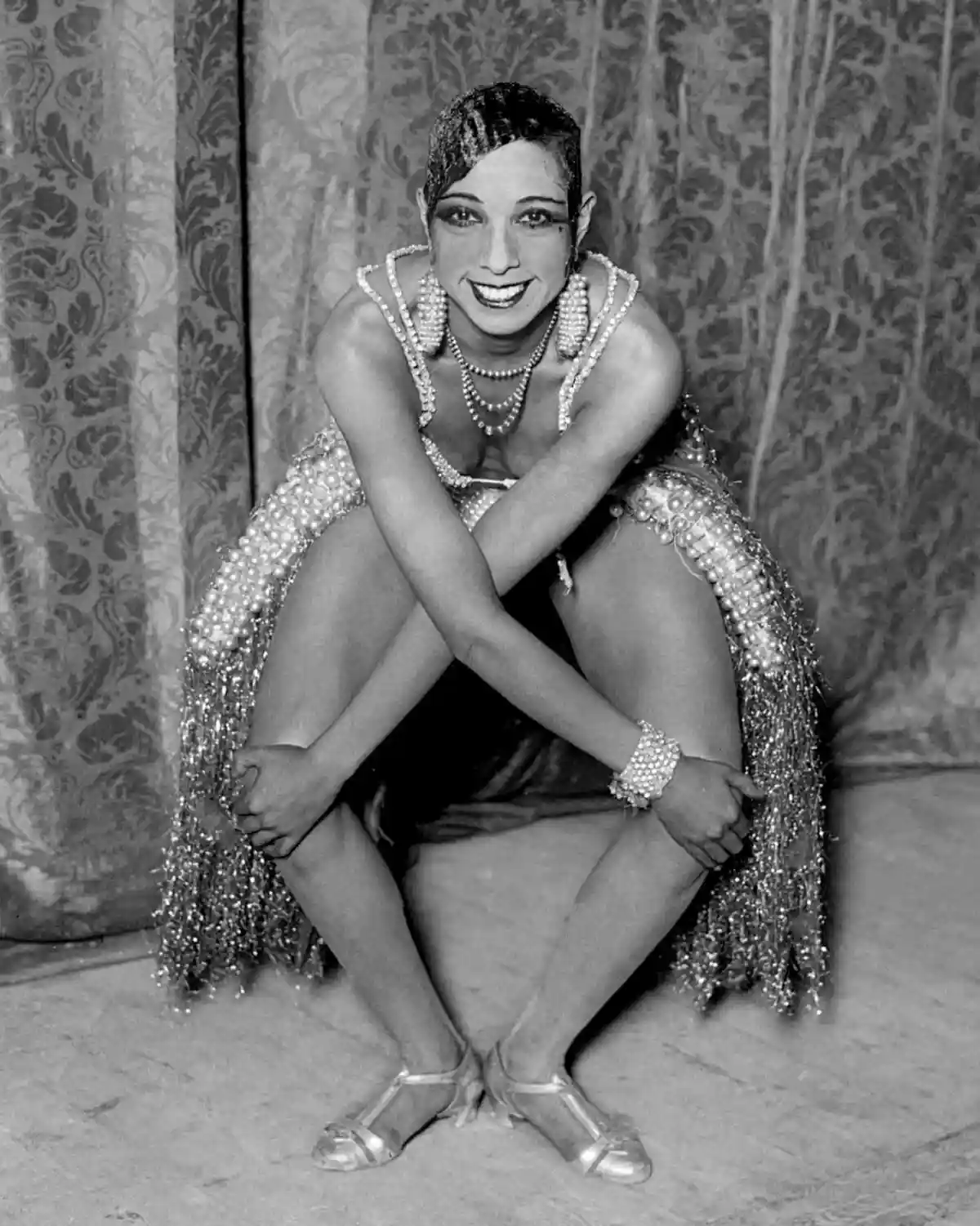 A vintage black-and-white photo shows a smiling woman in a pearl-encrusted flapper-style outfit crouching with her arms…