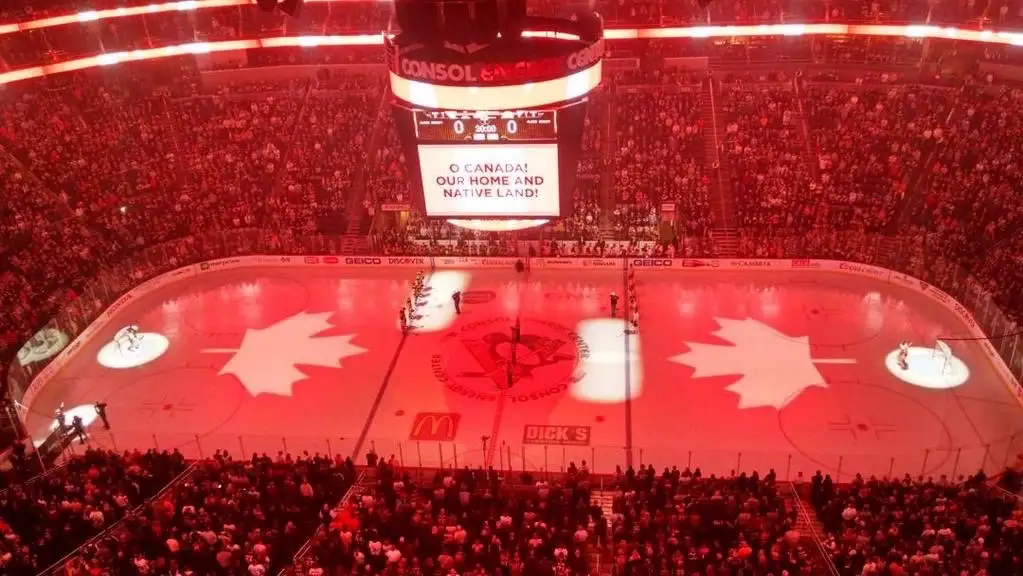 An overhead view of a packed ice hockey arena bathed in red light, with large maple leaf projections on the ice and a…