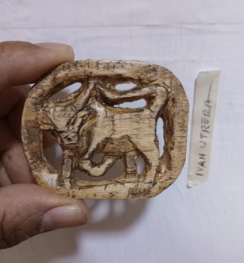 A hand holds a roughly carved wooden artifact depicting a stylized animal, possibly a bull or cow, with cut-out shapes…