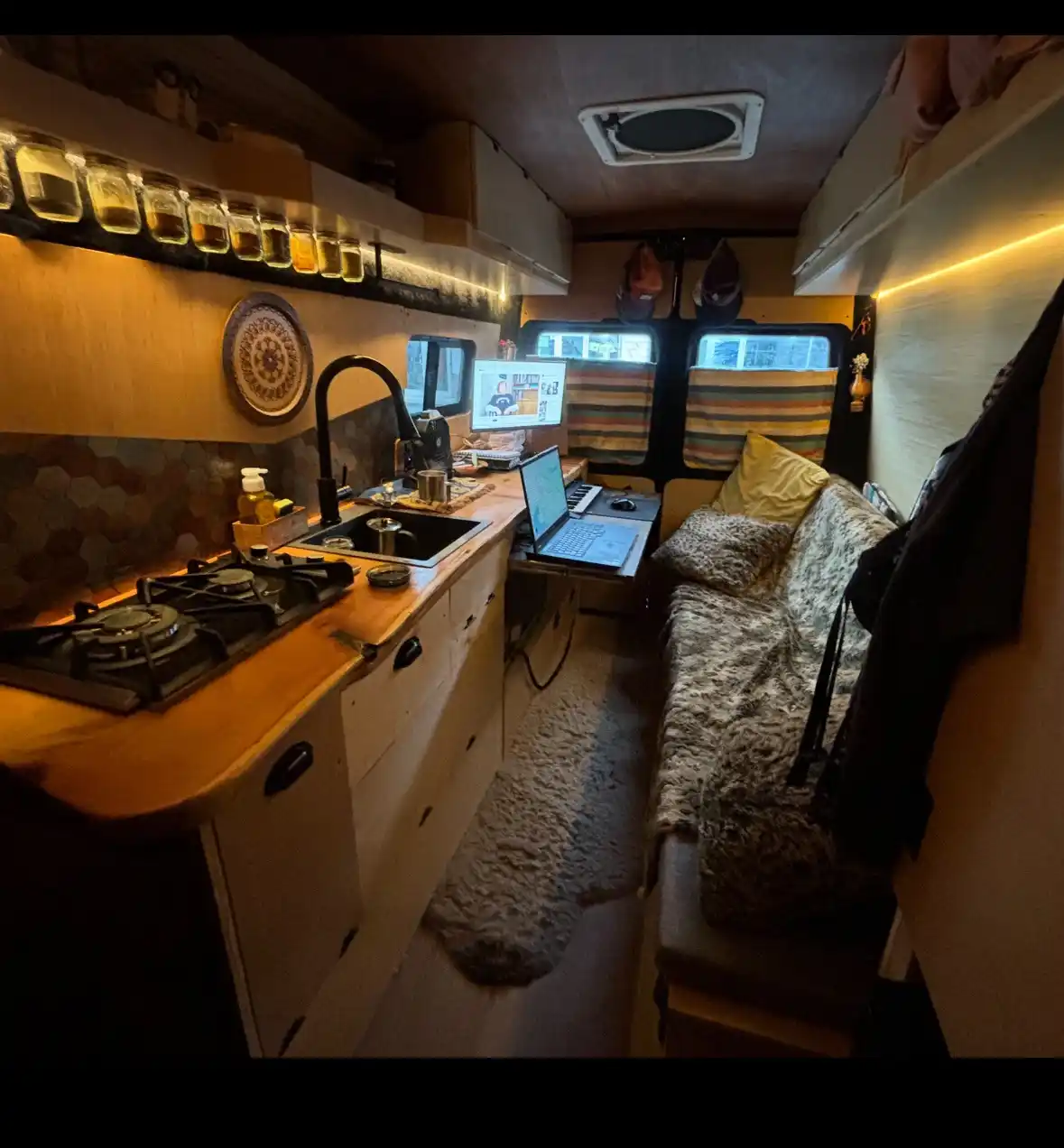 A cozy, compact van interior with a wooden countertop, sink, stove, and laptop on a small desk. A bed with a patterned…