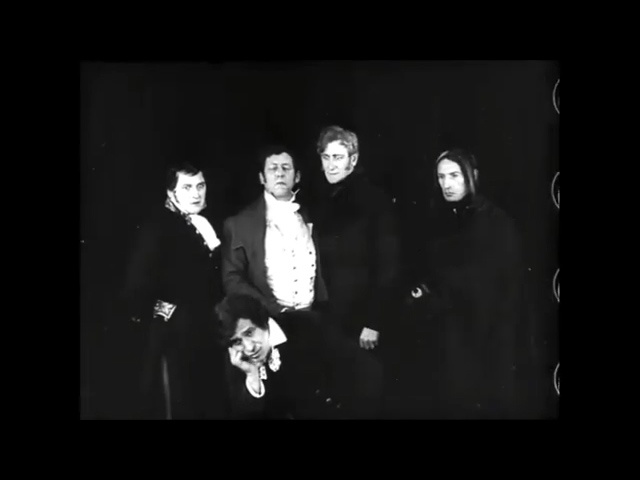 A black-and-white historical photograph shows five formally dressed individuals, four standing and one kneeling in front…