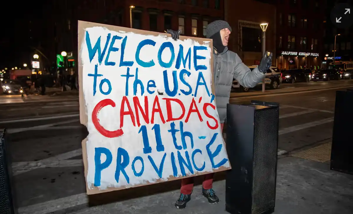 A person in winter clothing stands on a city sidewalk at night holding a large handmade sign that reads 'WELCOME to the…