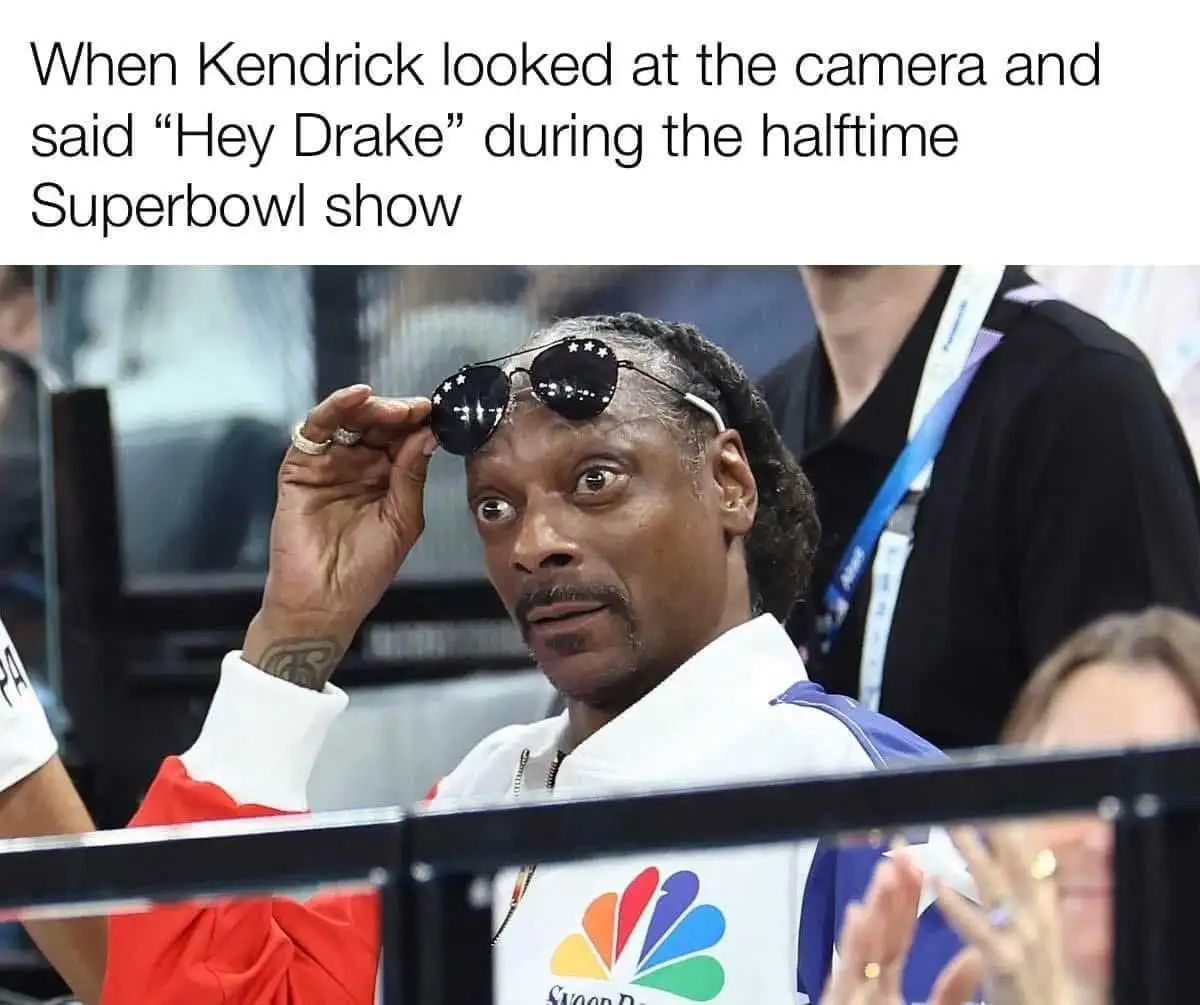 A man with braided hair and a white jacket is holding up sunglasses with stars on the lenses, looking surprised. He wears a…